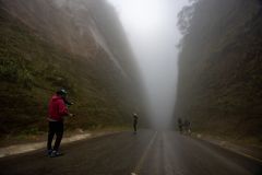 Grupo de pessoas caminha em rodovia entre duas montanhas, em meio a neblinas