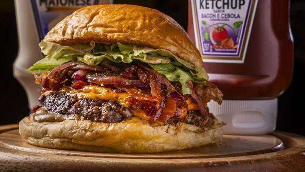 Hambúrguer do Mr. Mills criado para o evento com burger de 130g, queijo cheddar, cebola roxa assada e temperada, farofa de bacon, maionese Mills, ketchup Heinz Bacon e cebola caramelizada, alface americana picada no pão de brioche