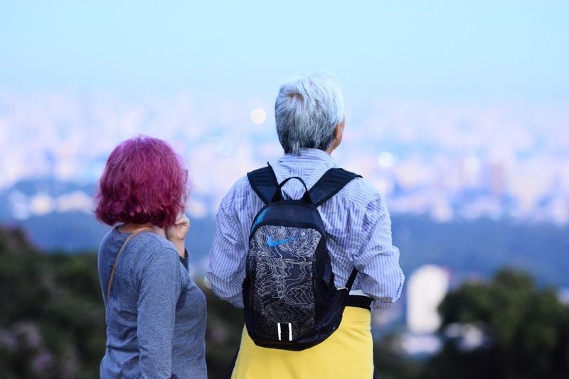 Duas mulheres de costas para a câmera realizando uma trilha pela natureza enquanto olham a paisagem