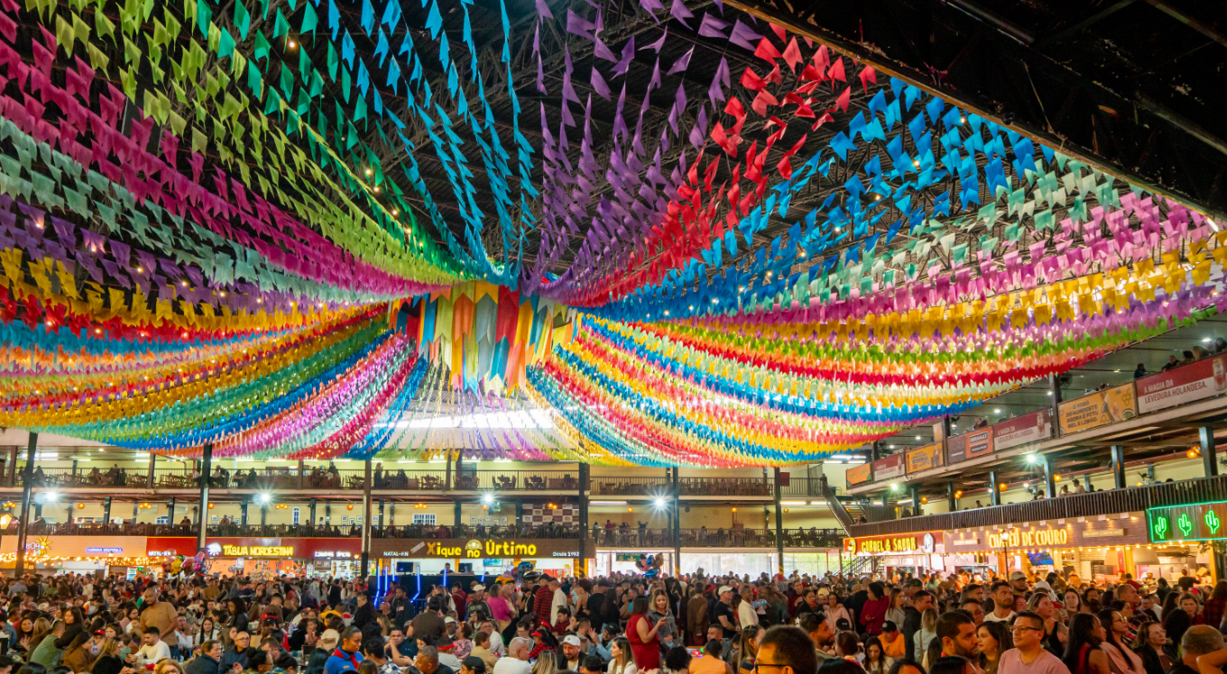 Imagem com bandeirinhas de Festa Junina coloridas e multidão em arraial